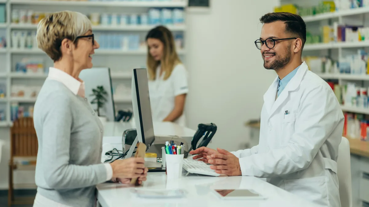 An older woman picking up a prescription from a pharmacist at a pharmacy counter, the kind of purchase covered by the state attorneys general generic drug settlements.